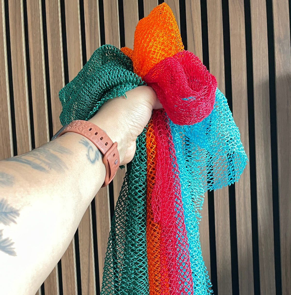 A close-up of a hand holding a multiple African Net sponges with colours teal green, orange, red, and turquoise blue against a dark wood slat background.