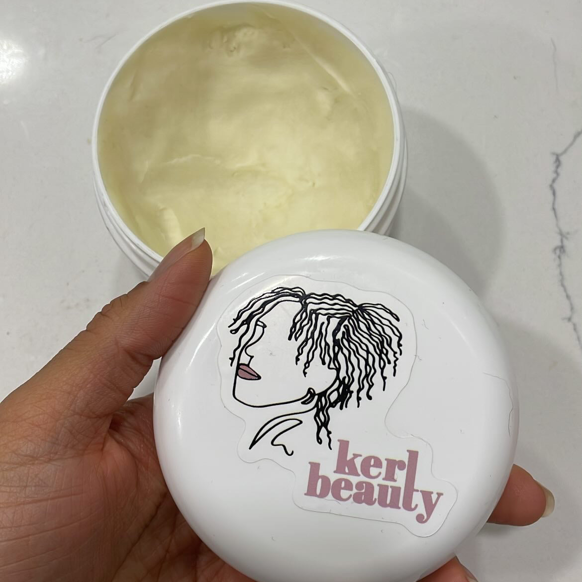 A hand holds a round white jar lid with a black line drawing of a woman's profile and the words 'kerl beauty' in pink, next to an open white jar containing pale yellow body butter, on a light marble surface.